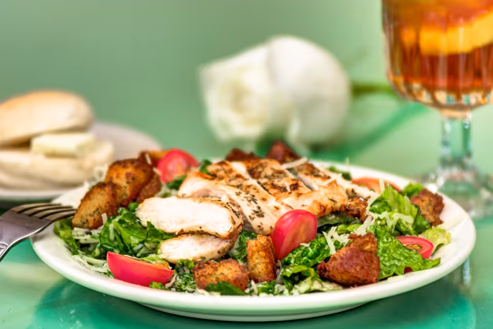 A plate of grilled chicken Caesar salad with romaine lettuce, cherry tomatoes, croutons, and shredded cheese. In the background, there is a glass of iced tea and a plate with two bread rolls and a pat of butter.