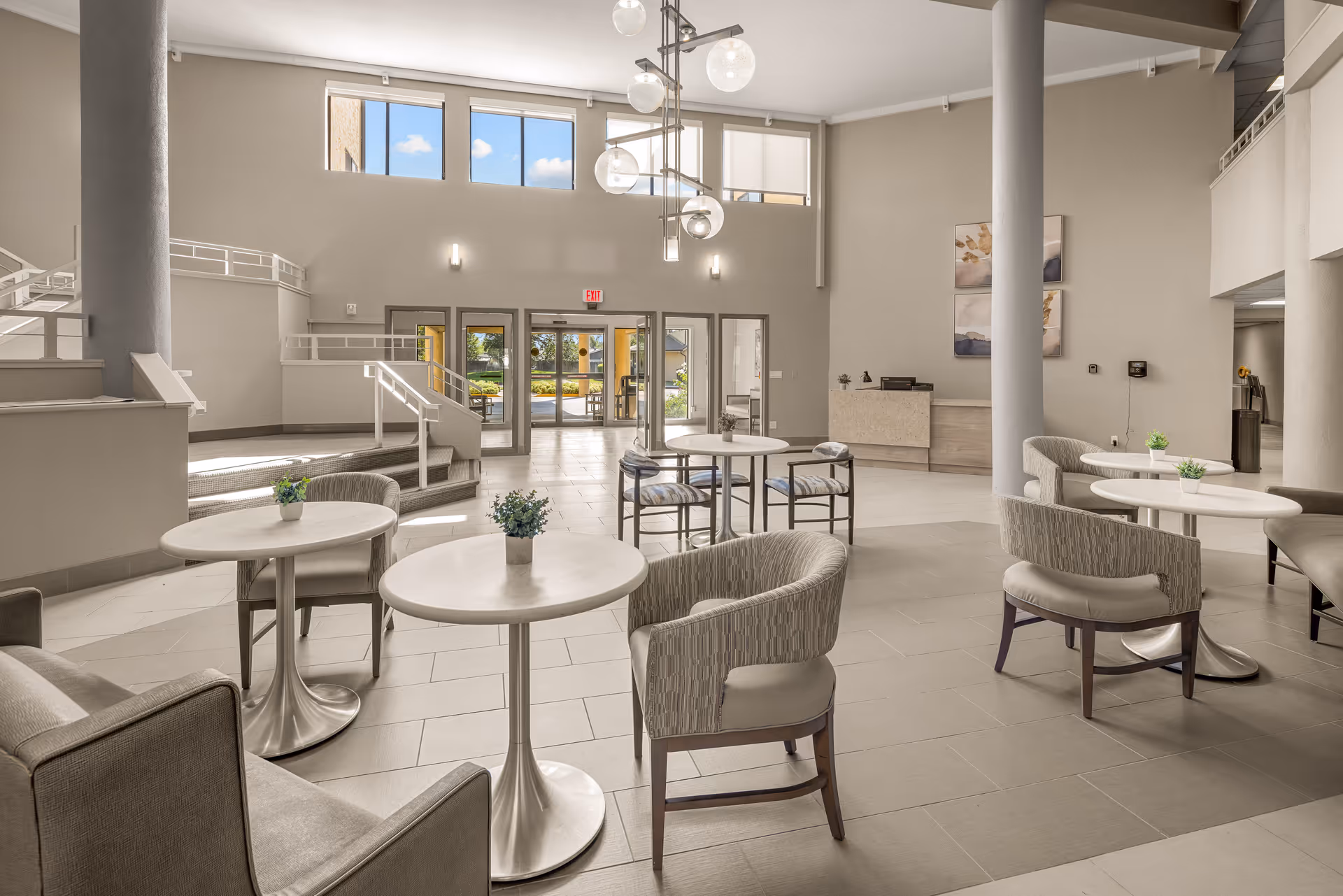 Spacious and bright senior living community common area with multiple round tables and cushioned chairs arranged for seating. Large windows near the ceiling allow natural light to fill the room. The entrance doors are visible in the background along with a reception desk and modern light fixtures hanging from the ceiling.