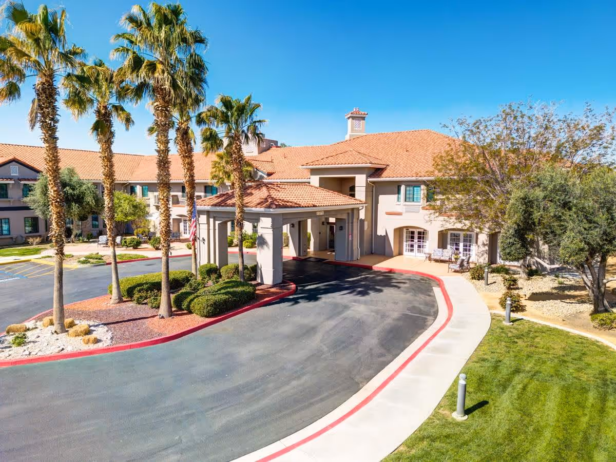 Exterior view of The Havens at Antelope Valley senior living facility featuring a driveway with a covered entrance, palm trees, landscaped bushes, and a clear blue sky.