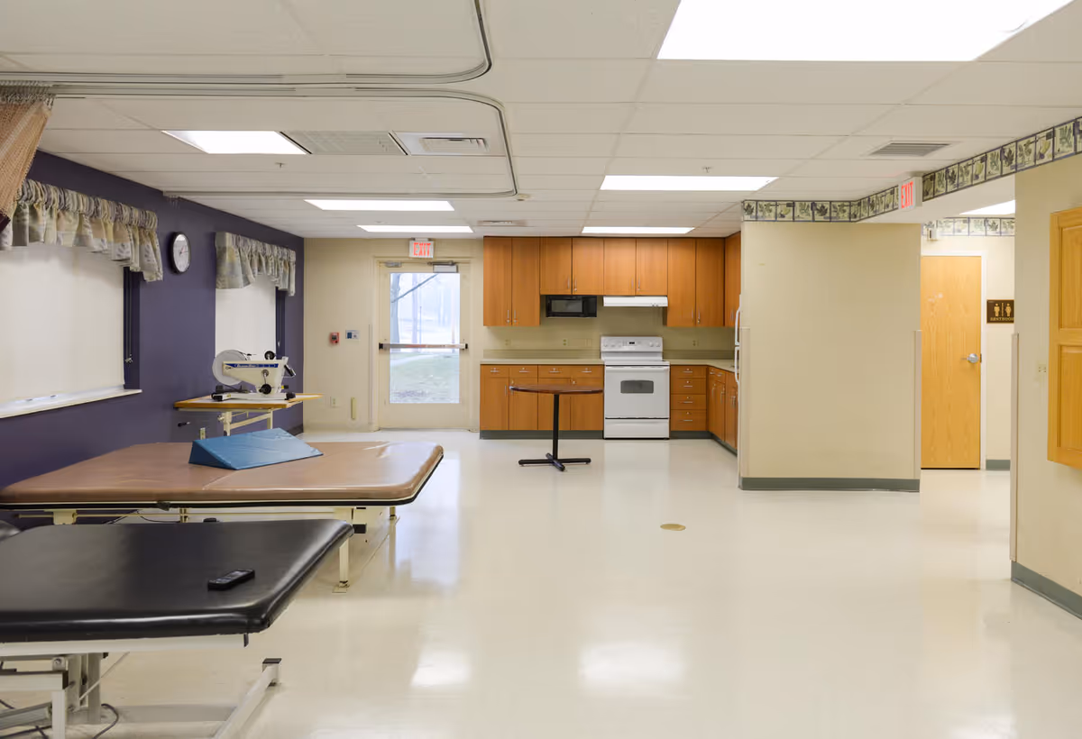 A rehabilitation room with two therapy tables, one brown and one black, positioned near a purple wall with windows covered by valances. There is a small table with a medical device on it. In the background, there is a kitchenette area with wooden cabinets, a white stove, microwave, and refrigerator. A door with an exit sign is visible, as well as a hallway leading to a restroom.