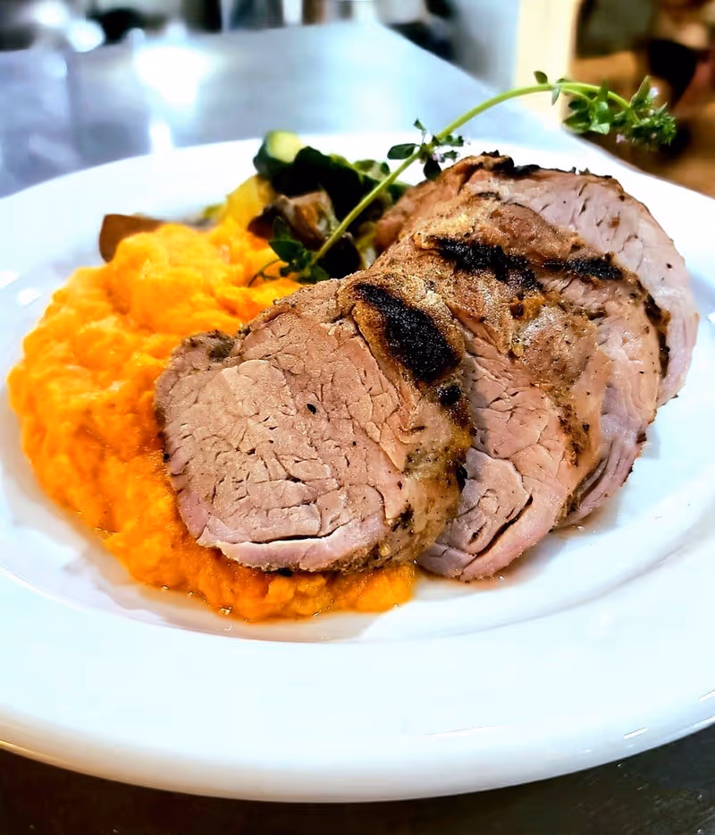 A white plate with slices of grilled pork tenderloin served on a bed of mashed sweet potatoes, garnished with a sprig of fresh herbs and some mixed vegetables in the background.