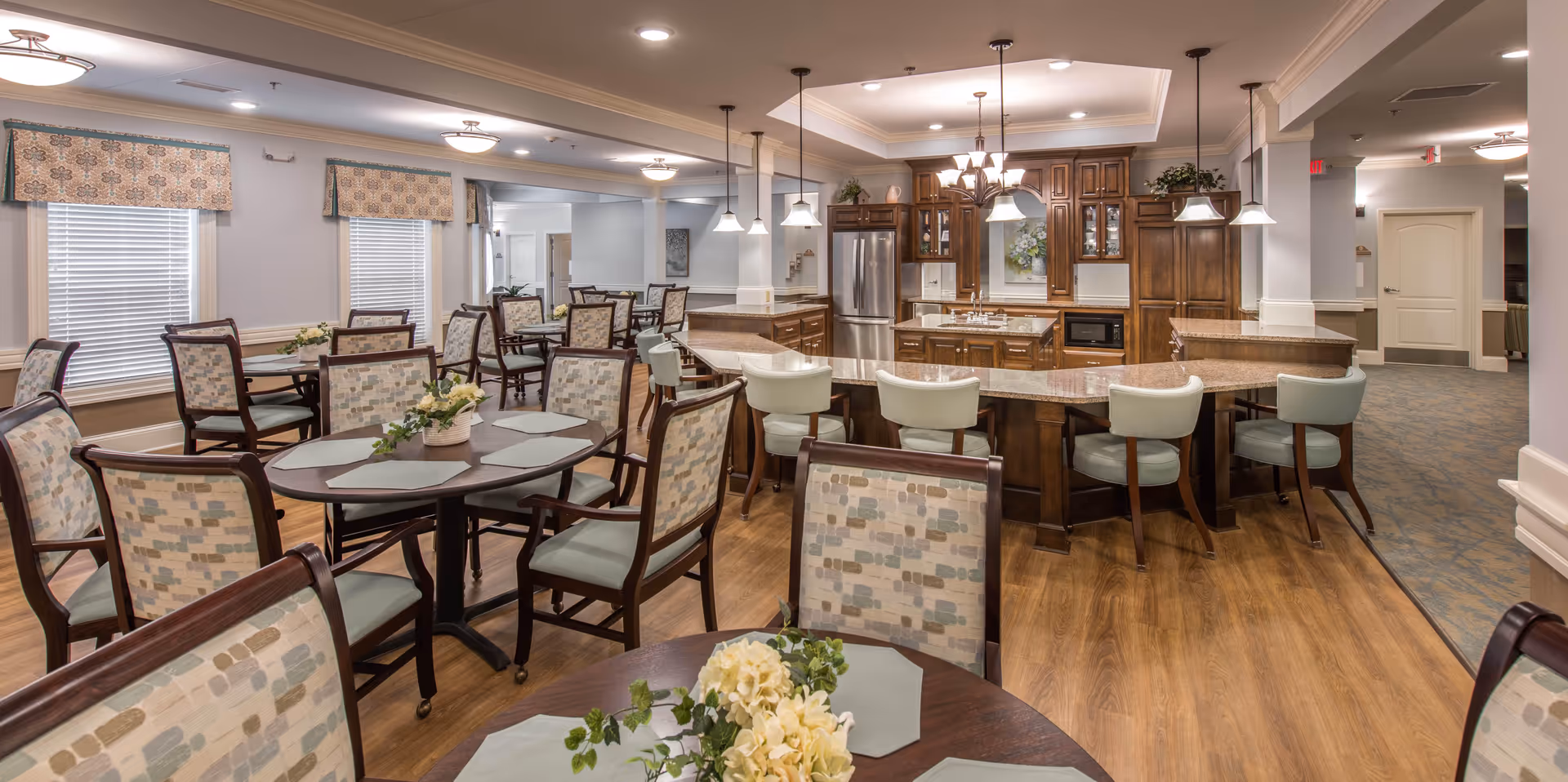 A spacious dining room in a senior living facility featuring multiple round tables with cushioned chairs arranged neatly. The room has large windows with patterned valances, wooden flooring, and a kitchen area with wooden cabinets, a refrigerator, and a curved countertop with bar stools. Soft lighting fixtures hang from the ceiling, creating a warm and inviting atmosphere.
