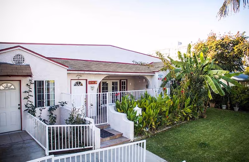 Exterior view of a single-story residential building with white walls and a brown roof. The entrance features a white door and a small porch area with white railings. There are green plants and bushes along the front of the building and a well-maintained lawn to the right. Trees and other greenery are visible in the background under a clear sky.