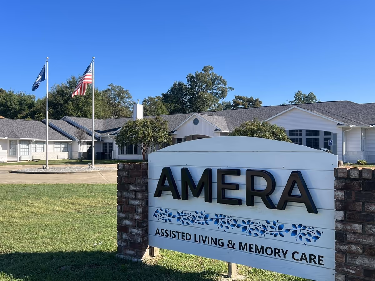 Exterior view of Amera Assisted Living & Memory Care facility with a large sign in the foreground displaying the facility name. The building is single-story with white siding and a gray roof. Two flagpoles with the American flag and another flag are visible on the left side, along with green grass and trees under a clear blue sky.