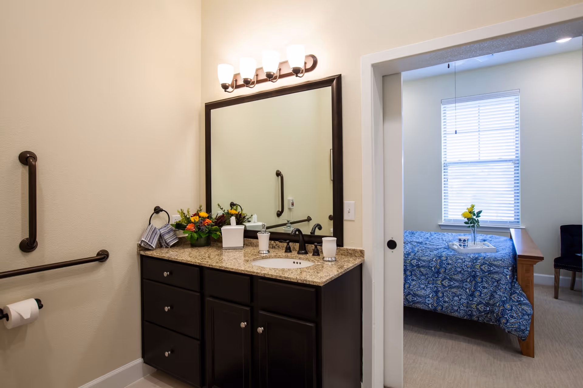Bathroom vanity with a large mirror, granite countertop, dark wood cabinets, and a light fixture with five lights above. There are handrails on the wall and a toilet paper holder. Through an open doorway, a bedroom with a bed covered in a blue patterned bedspread and a window with blinds is visible.