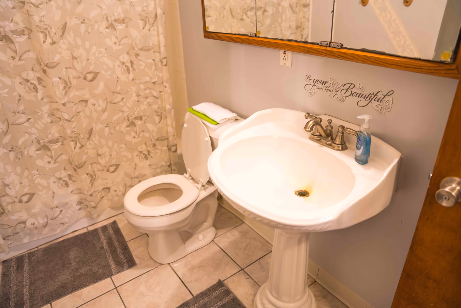 Small bathroom with a pedestal sink, toilet, leaf-patterned shower curtain, and mirrored medicine cabinet.