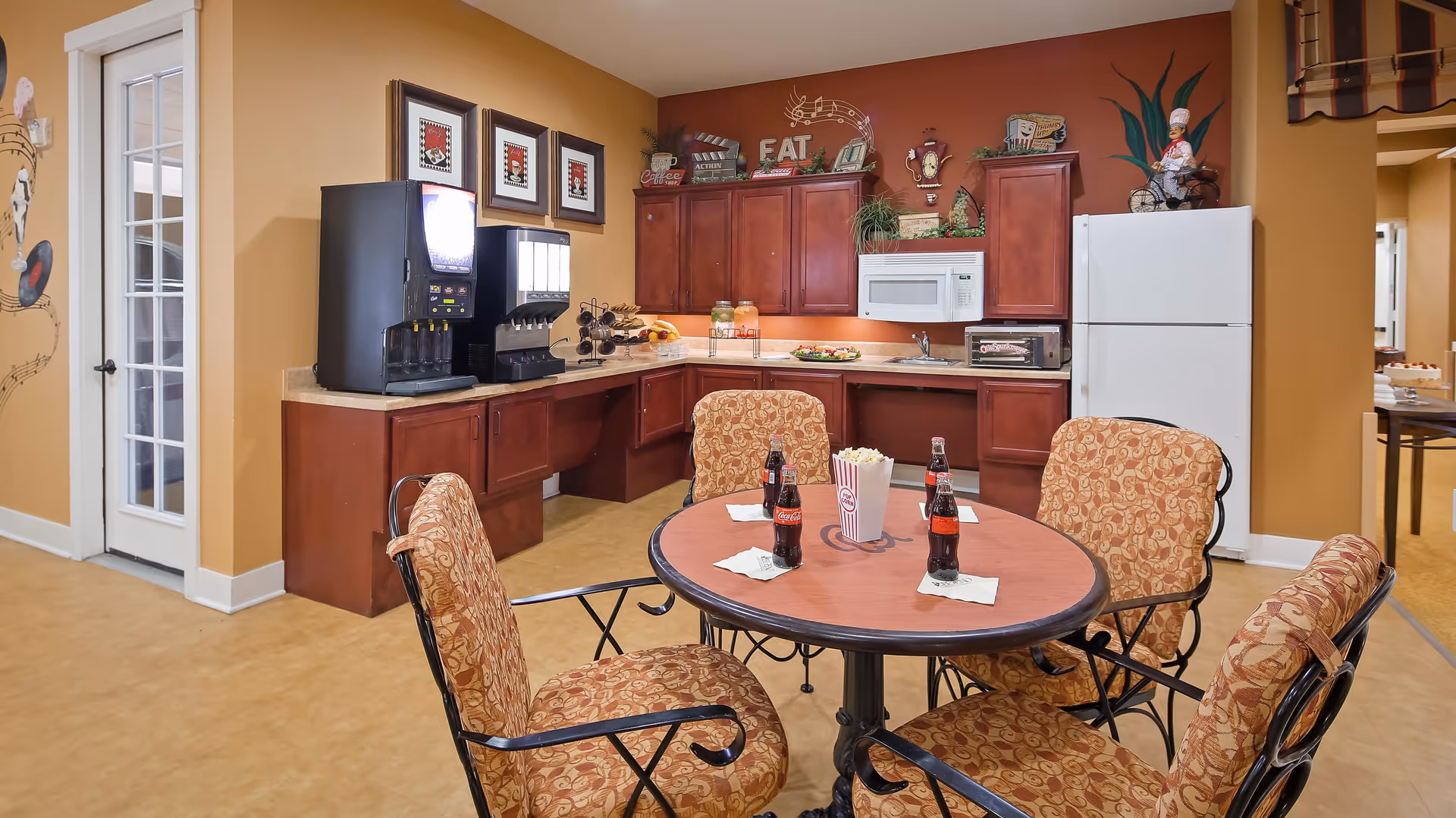 A cozy kitchen and dining area with a round table surrounded by four cushioned chairs. On the table are three bottles of Coca-Cola and a container of popcorn. The kitchen features wooden cabinets, a white microwave, a white refrigerator, a toaster oven, and beverage dispensers. The walls are painted warm tones with decorative items and framed pictures.