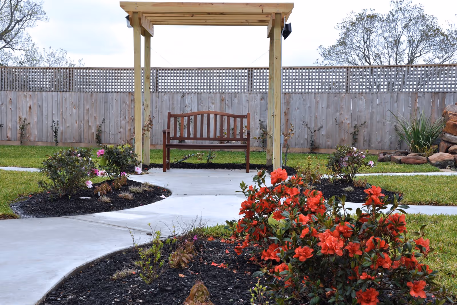 A peaceful outdoor garden area with a wooden bench under a wooden pergola. The garden features a concrete pathway winding through flower beds with red and pink blooming flowers, surrounded by green grass and a wooden fence in the background.