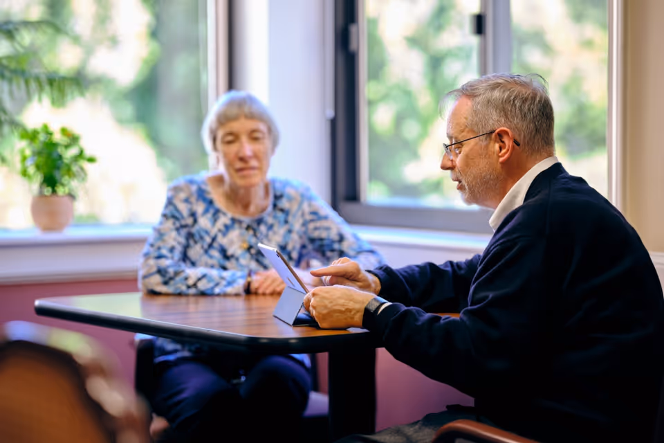 An elderly man and woman sitting at a wooden table in a room with large windows. The man is using a tablet device while the woman watches attentively. There is a potted plant on the windowsill and greenery visible outside the window.
