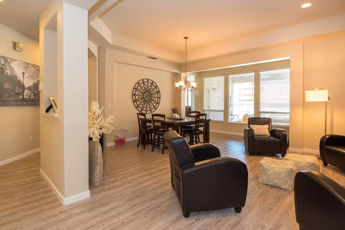 A bright and spacious senior living common area featuring a dining table with six chairs, three black leather armchairs arranged around two patterned ottomans, a floor lamp, and large windows with blinds allowing natural light to fill the room. The walls are painted beige and decorated with a large circular metal wall art and a framed black and white photograph.