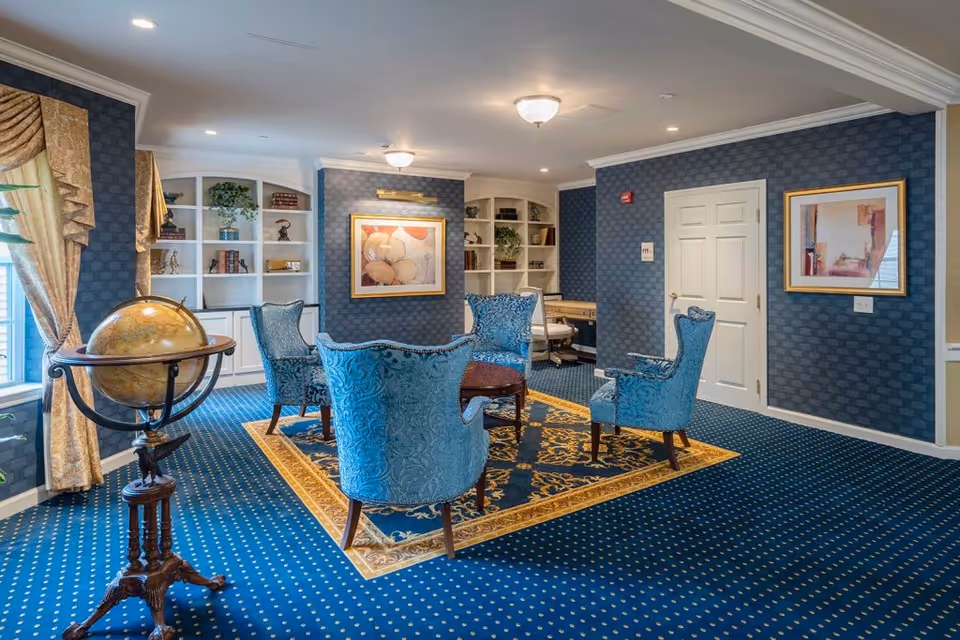 A cozy sitting area in a senior living facility with four blue upholstered armchairs arranged around a small wooden table on a decorative blue and gold rug. The room features blue patterned carpet and wallpaper, white built-in shelves with books and decorative items, framed artwork on the walls, a globe on a wooden stand near a window with gold curtains, and a desk with a chair in the background.