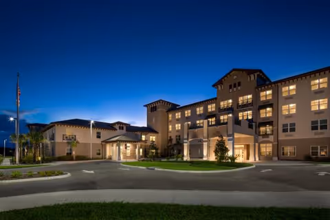 Exterior view of a large, well-lit senior living facility building at dusk with multiple windows and a covered entrance. The sky is clear and dark blue, and there is a circular driveway with landscaped greenery in front.