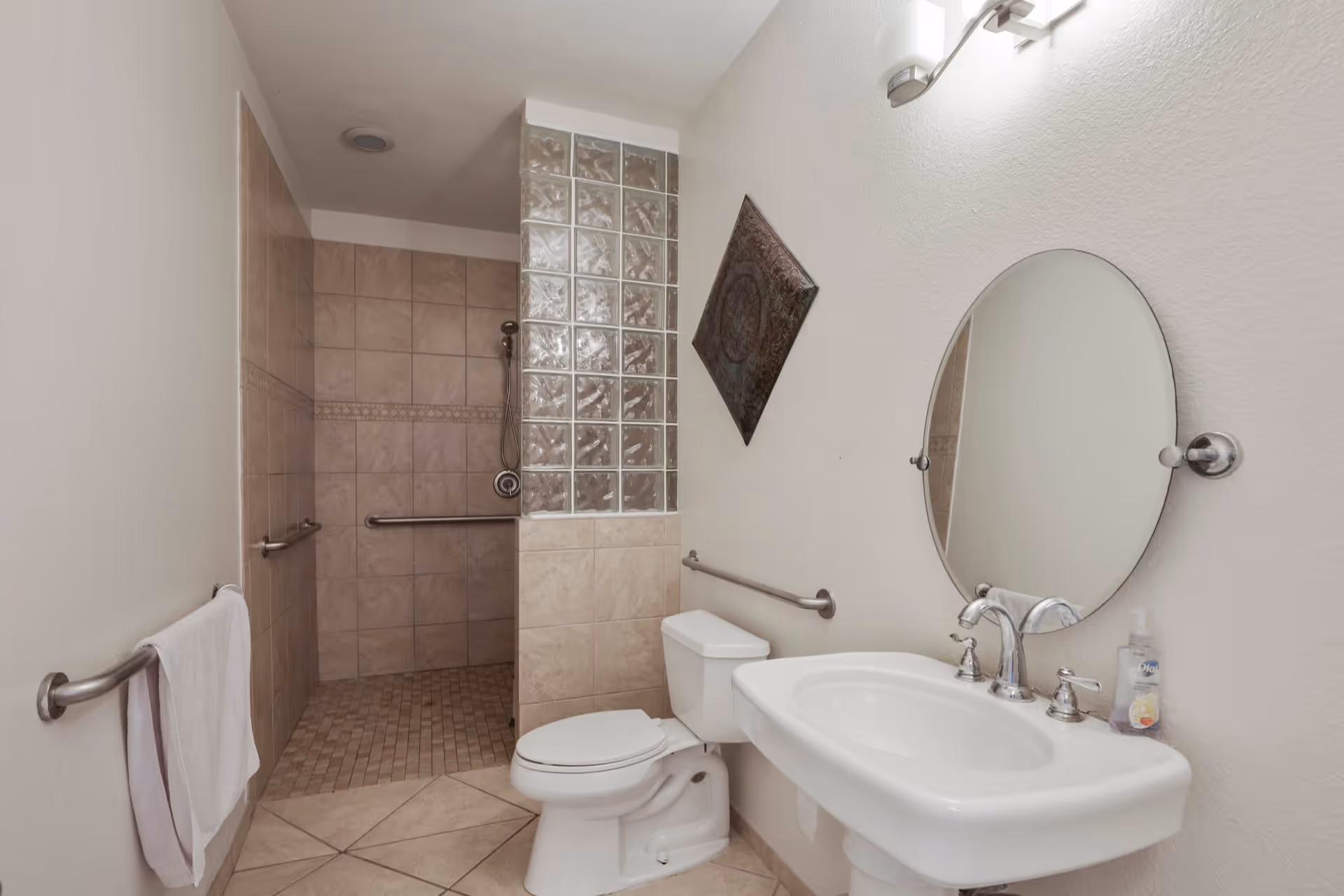 A clean bathroom featuring a walk-in tiled shower with grab bars, a white toilet, a white sink with chrome fixtures, an oval mirror above the sink, a towel hanging on a grab bar, and a decorative wall piece. The shower has a partial glass block wall.