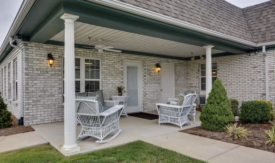 Covered outdoor patio area at a senior living facility with white wicker rocking chairs and a small table, surrounded by brick walls and greenery including bushes and a small tree.