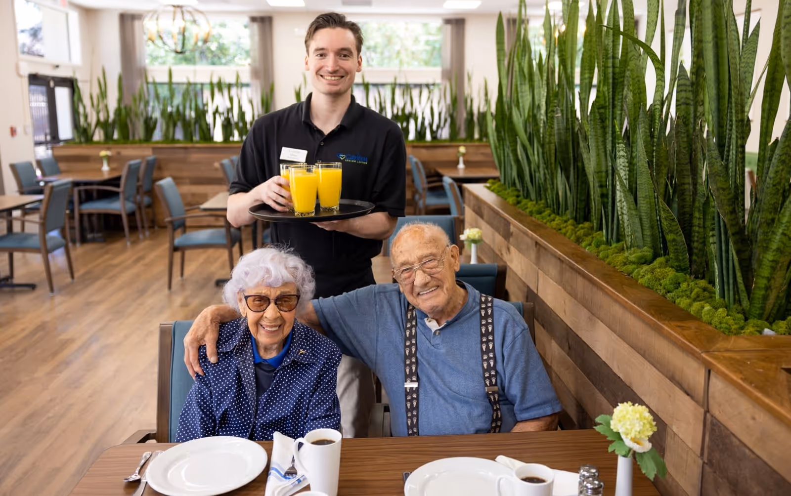 A smiling male staff member holding a tray with four glasses of orange juice stands behind an elderly couple seated at a dining table in a bright dining room with wooden floors and green plants along the wall. The elderly woman wears glasses and a blue polka dot shirt, and the elderly man wears glasses, a blue shirt, and suspenders. The table is set with white plates, coffee cups, and silverware.
