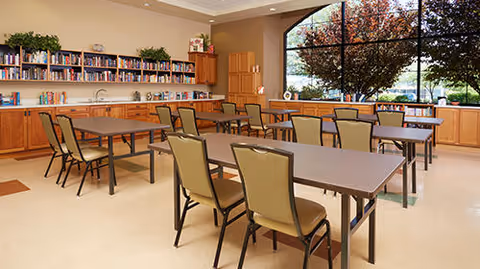 A spacious dining area with multiple tables and chairs arranged neatly. The room features large windows with a view of trees outside, wooden cabinets along the walls, and a bookshelf filled with books. The space is well-lit with natural light.
