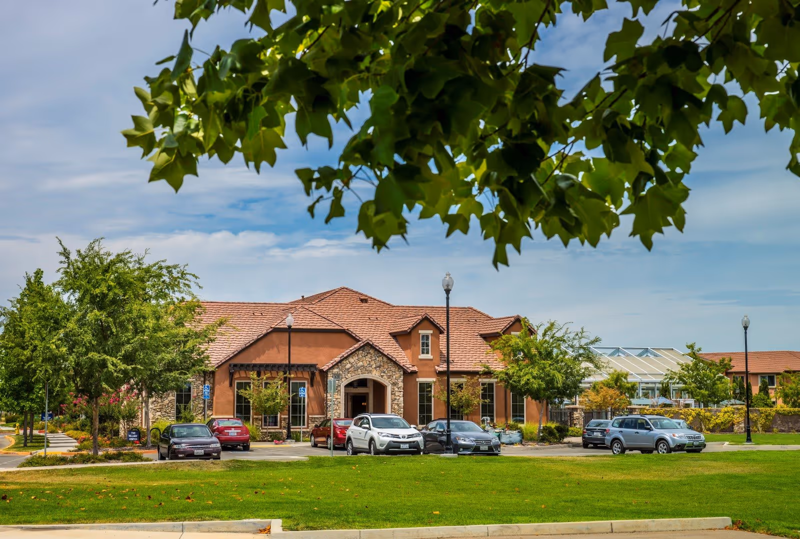 Exterior view of Eskaton Village Roseville building with a stone and stucco facade, red tiled roof, several parked cars in front, green lawn, trees, and a partly cloudy sky.