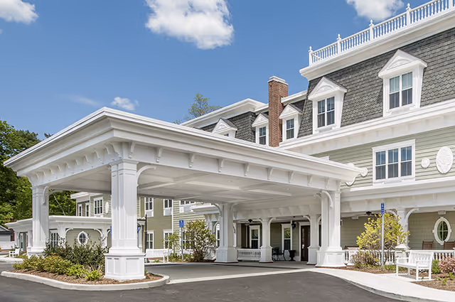 Exterior view of Brightview on New Canaan senior assisted living and memory care facility showing a large covered entrance with white columns, a paved driveway, landscaped bushes, and a multi-story building with dormer windows under a blue sky with a few clouds.