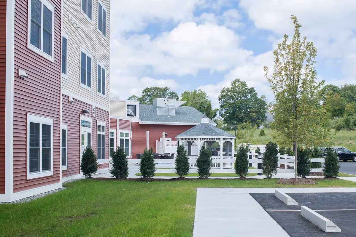 Exterior view of Elmbrook Village At Bozrah/Norwich showing a red and white multi-story building with multiple windows, a white gazebo surrounded by small bushes, a tree, green grass, and a parking area under a partly cloudy sky.