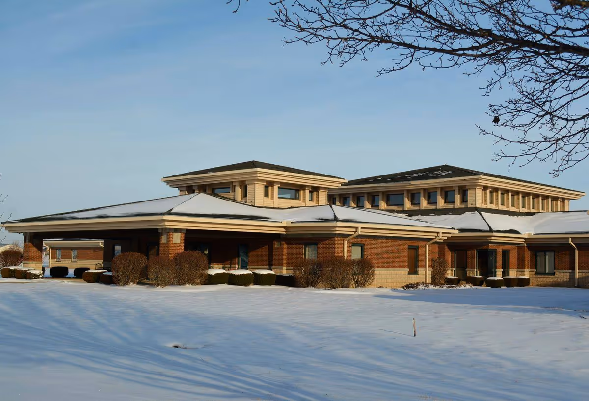 Exterior view of a single-story brick building with a snow-covered roof and surrounding snowy ground under a clear blue sky. The building has multiple windows and some bushes around it.