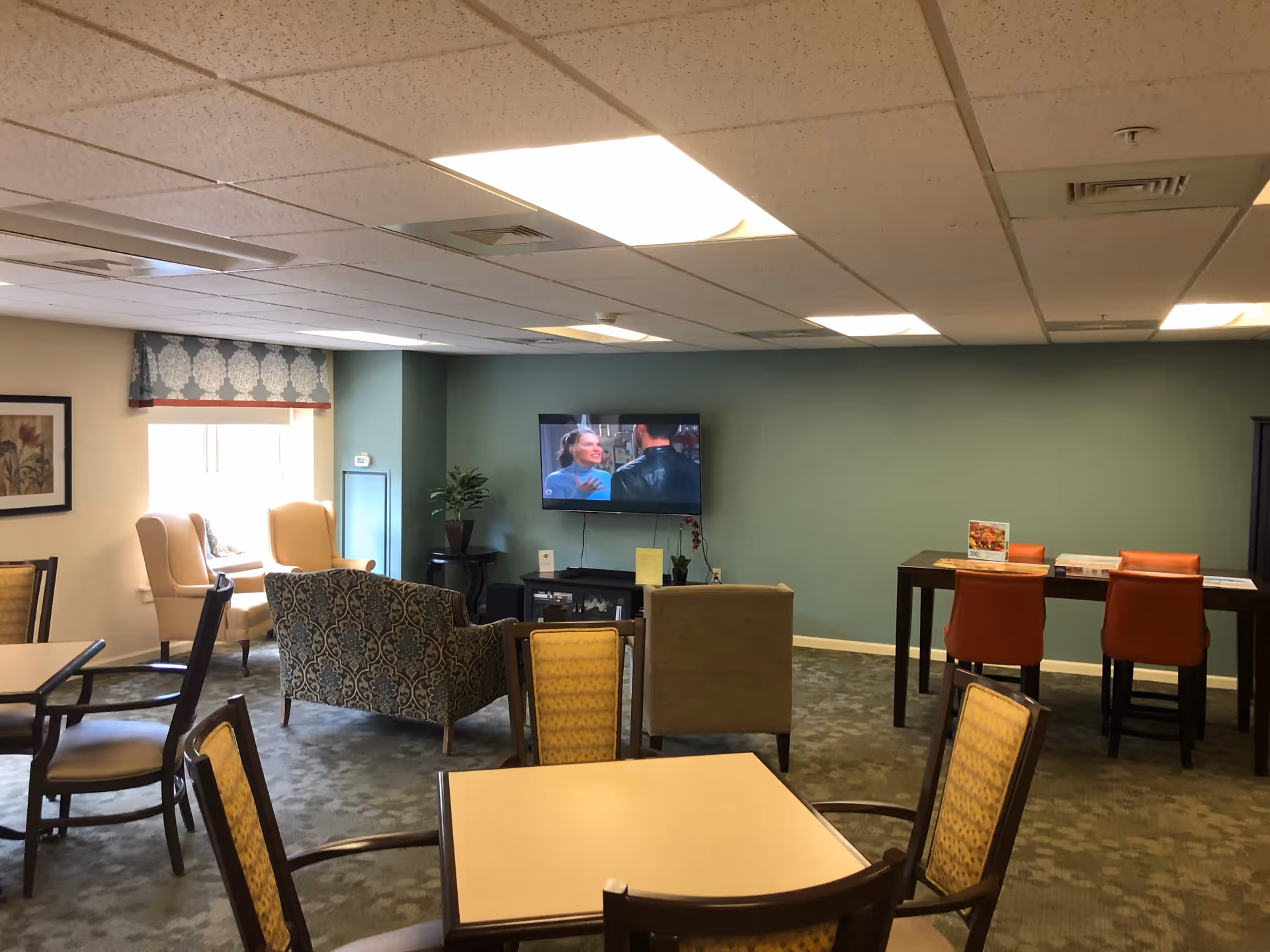 A senior living facility common area with multiple seating options including armchairs, a patterned loveseat, and dining tables with chairs. A flat-screen TV is mounted on a green wall, showing a scene with two people talking. There is a window with a decorative valance, a potted plant, and a high table with orange chairs and puzzles on top.