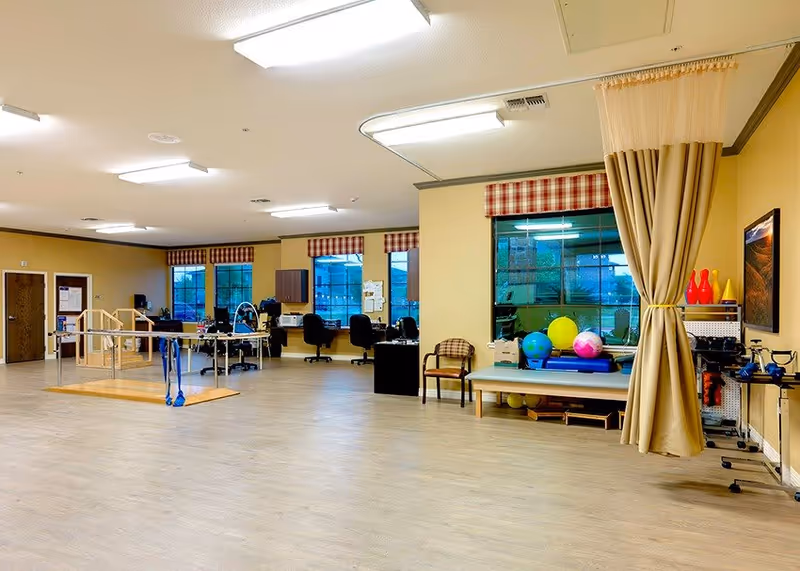 A spacious physical therapy or rehabilitation room with wooden handrails for walking exercises, several exercise balls, therapy equipment, chairs, desks with office chairs, and large windows with red and white checkered valances. The room has light-colored wood flooring and beige walls with a curtain partially drawn to one side.