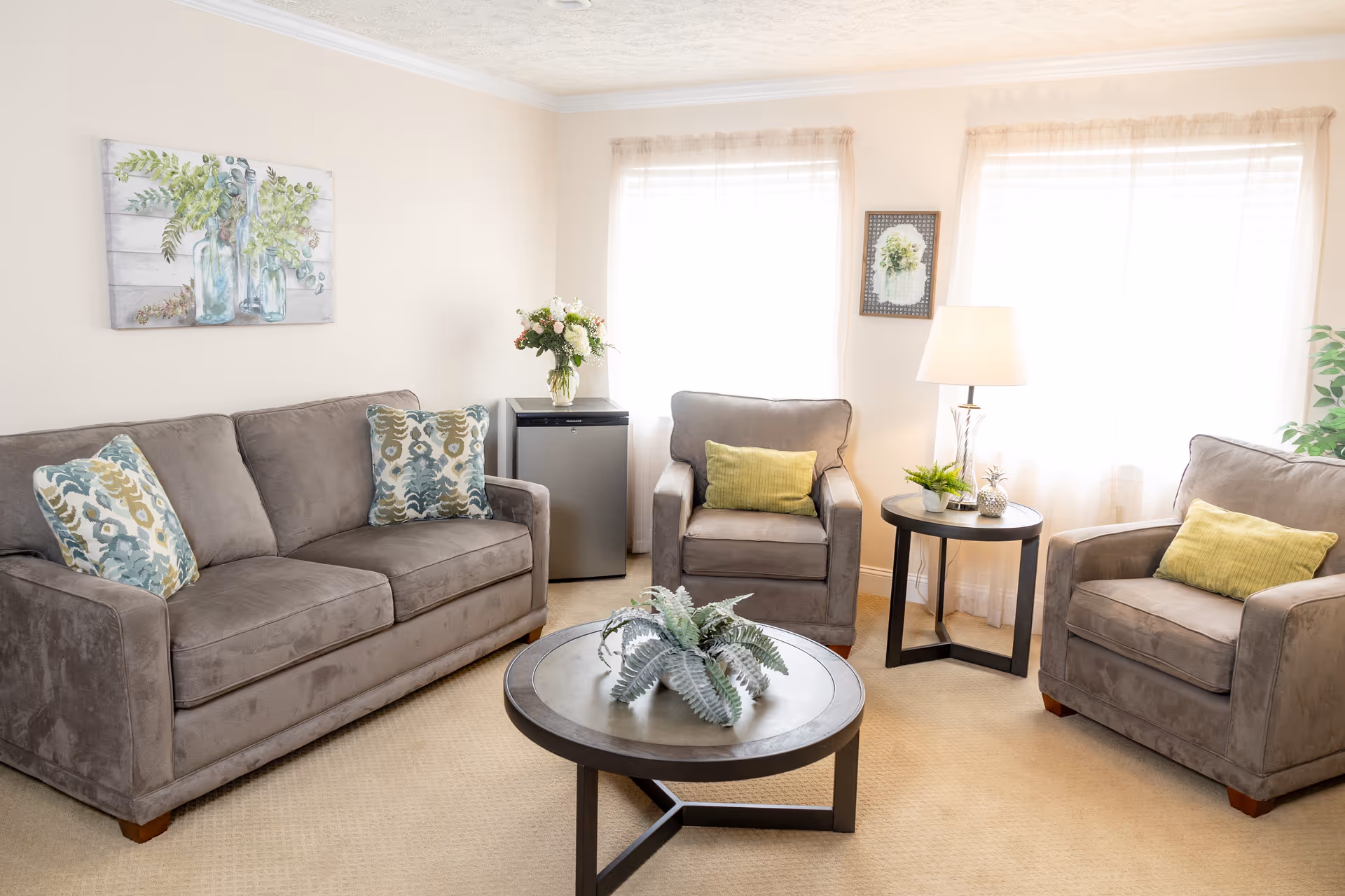 A bright living room with a gray sofa and two matching armchairs, each with decorative pillows. There is a round coffee table with a plant centerpiece in the middle, a side table with a lamp and small plants, and a small refrigerator in the corner. The walls are light-colored with two windows covered by sheer curtains, and a floral painting and framed artwork hang on the walls.