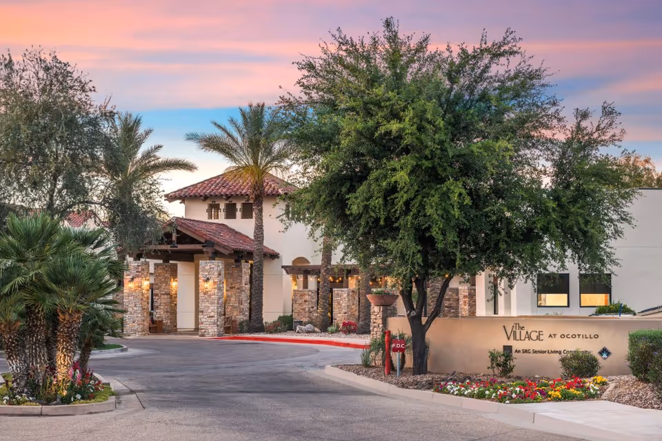 Exterior view of The Village at Ocotillo senior living facility during sunset, featuring a driveway lined with palm trees and other greenery, stone pillars at the entrance, and a sign with the facility's name surrounded by colorful flowers.