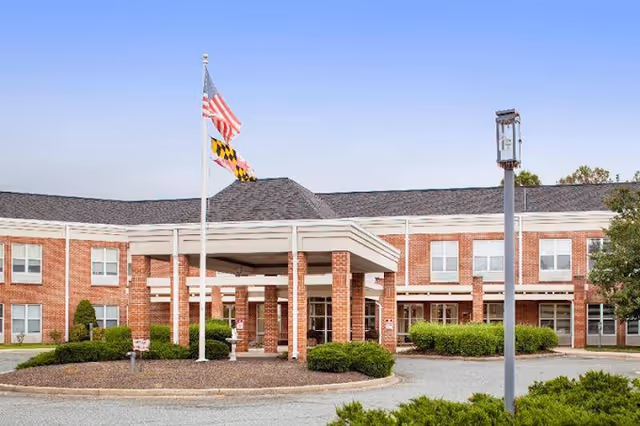 Exterior view of a two-story brick nursing and rehabilitation facility with a covered entrance supported by brick columns. Two flagpoles display the American flag and the Maryland state flag. There are shrubs and small trees around the building and a clear sky above.