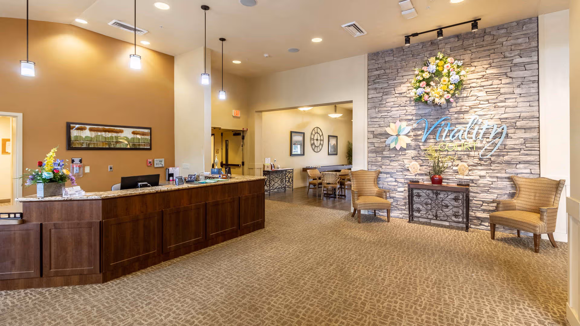 Reception area of Vitality Court featuring a wooden front desk with a granite countertop, pendant lights hanging from the ceiling, a stone accent wall with the Vitality Court logo and a floral wreath above it, two upholstered chairs, and a small table with a vase of flowers. In the background, there is a seating area with tables and chairs and framed wall art.