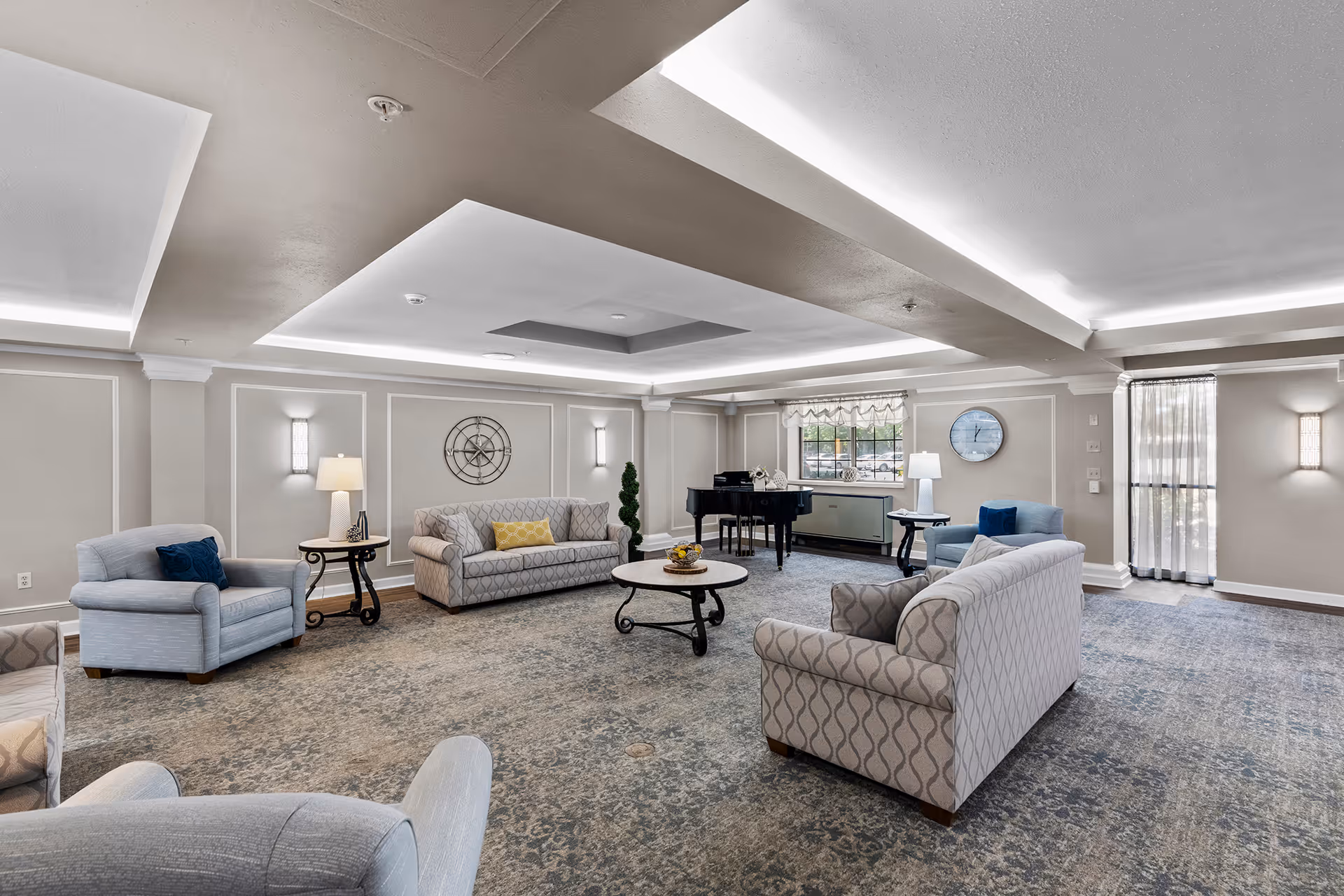 A spacious and well-lit living room area with multiple upholstered armchairs and sofas arranged around a central round coffee table. The room features soft carpeting, wall sconces, table lamps, a large window with sheer curtains, a wall clock, and a black grand piano in the corner. The ceiling has recessed lighting and decorative molding.