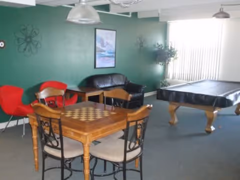 Community lounge with a chessboard game table, chairs, a couch, and a pool table in front of a green wall and window blinds.