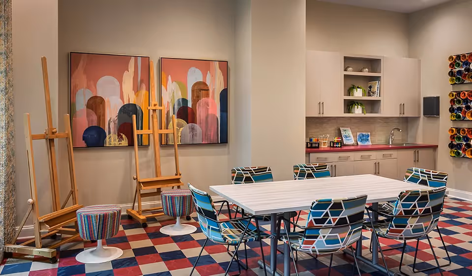 A colorful art room with a white table surrounded by six patterned chairs. The room features three wooden easels, two abstract paintings on the wall, a kitchenette with cabinets and a sink, and a wall-mounted organizer holding multiple rolls of colored paper. The floor has a red, blue, and beige checkered pattern.