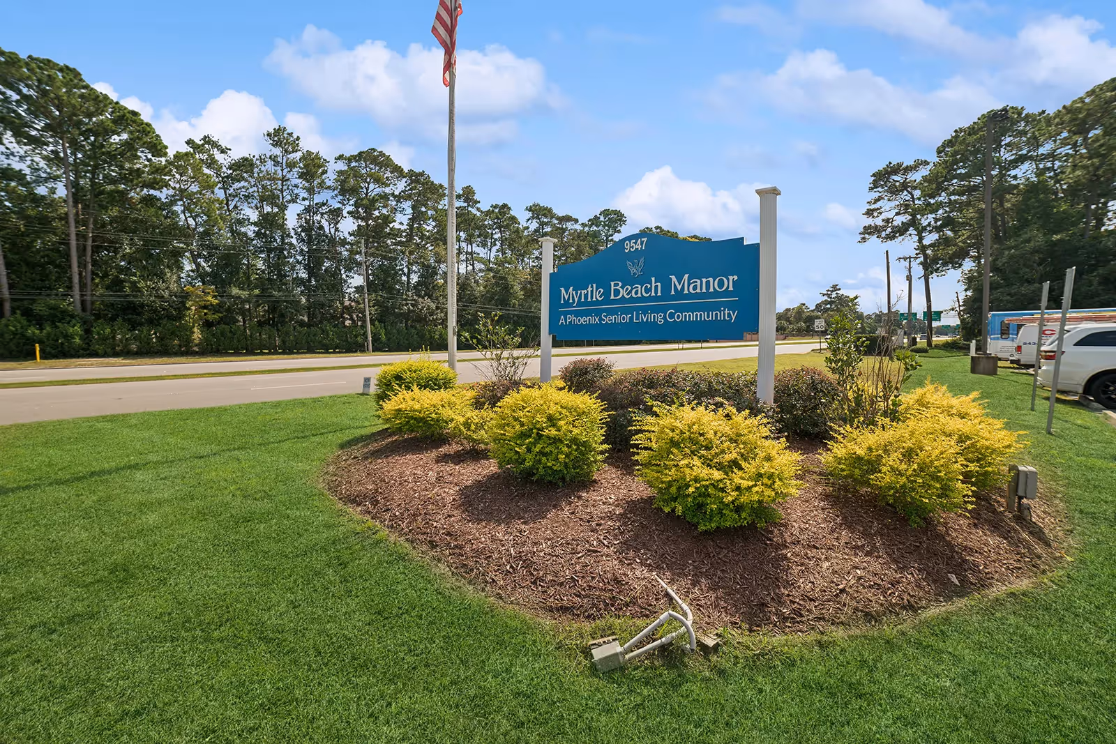 Outdoor view of a landscaped area with green grass and bushes surrounding a blue sign that reads 'Myrtle Beach Manor, A Phoenix Senior Living Community' with an American flag on a pole behind the sign and a road and trees in the background.