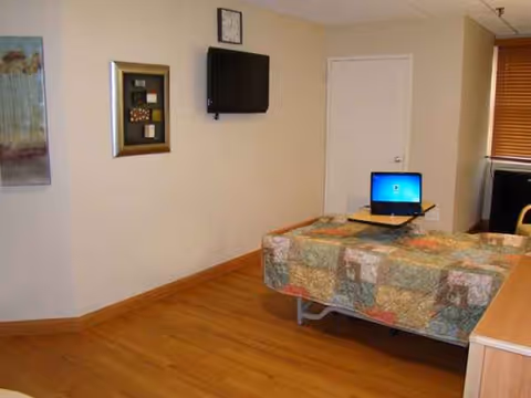 Interior of a senior living room/bedroom with wall-mounted TV, framed art, door, window, and bed.