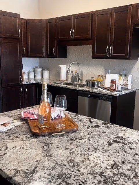 Modern kitchen with dark wooden cabinets, granite countertops, a stainless steel dishwasher, and a sink with a curved faucet. On the countertop, there is a wooden tray holding a bottle of rosé wine and two wine glasses, along with a folded pink and white cloth. Various kitchen items and containers are placed near the backsplash.
