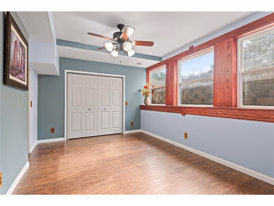 Empty room with light blue walls, wooden floor, three large windows with wooden frames, a ceiling fan with lights, a white double-door closet, and a framed picture on the left wall.