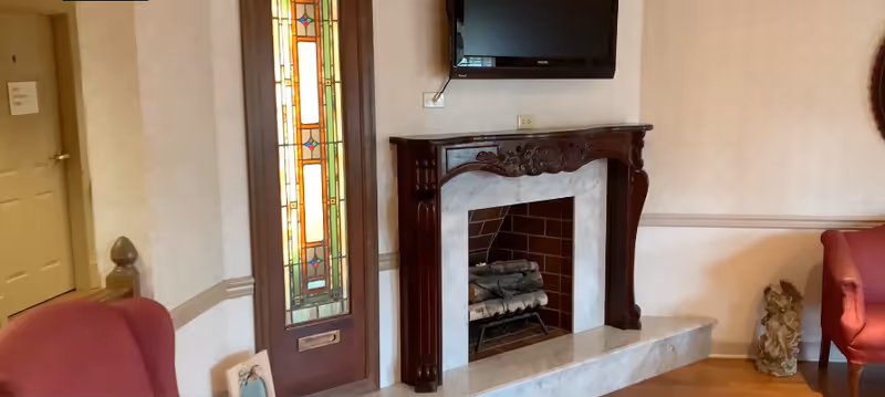 A cozy living room area featuring a decorative fireplace with a dark wooden mantel and a marble surround. Above the fireplace is a mounted flat-screen TV. To the left of the fireplace is a tall stained glass window with colorful geometric patterns. There are two red upholstered chairs partially visible on either side of the fireplace. The room has light-colored walls with a chair rail and wooden flooring.