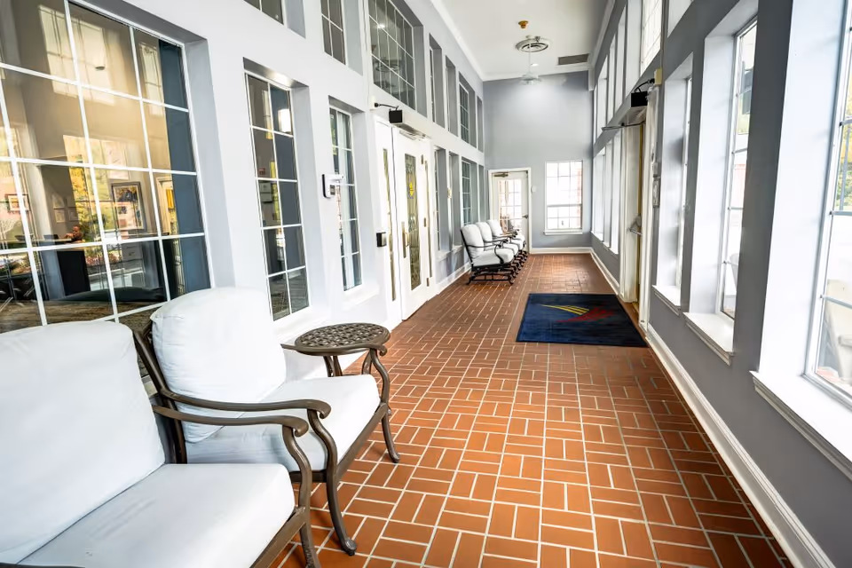 A bright indoor hallway with large windows on one side and glass-paneled doors on the other. The floor is covered with reddish-brown tiles arranged in a herringbone pattern. Several cushioned chairs with white upholstery and dark wooden frames are placed along the hallway, accompanied by small round tables. A dark blue mat with a colorful logo is near the door at the far end.