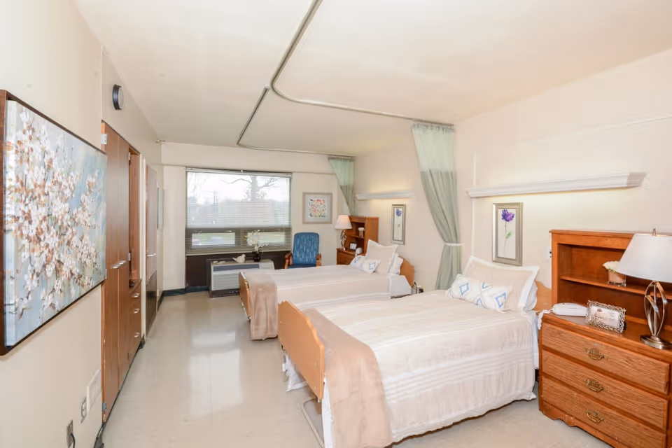 A bright and clean room with two single beds, each with beige bedding and pillows. There is a wooden nightstand with a lamp and a framed photo between the beds. The room has a large window with blinds, a blue armchair, and floral artwork on the walls. Light green curtains hang on the walls behind the beds.