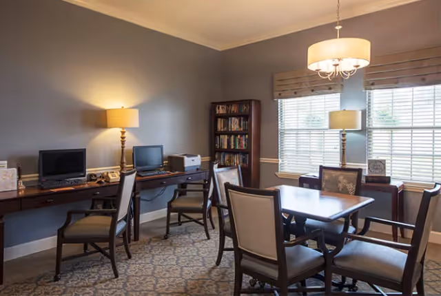 A well-lit room with a table and four chairs in the center, two desks with computers and lamps along the walls, a bookshelf filled with books, and two large windows with blinds letting in natural light.