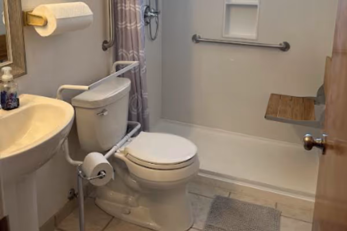 A small bathroom with a white toilet equipped with safety rails, a pedestal sink with a soap dispenser, a shower area with a wall-mounted wooden seat and grab bars, and a shower curtain with a circular pattern.