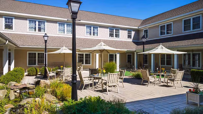 Outdoor courtyard area of a senior living facility with patio tables, chairs, umbrellas, lampposts, and landscaped greenery including bushes and a small rock water feature, surrounded by a two-story building under a clear blue sky.