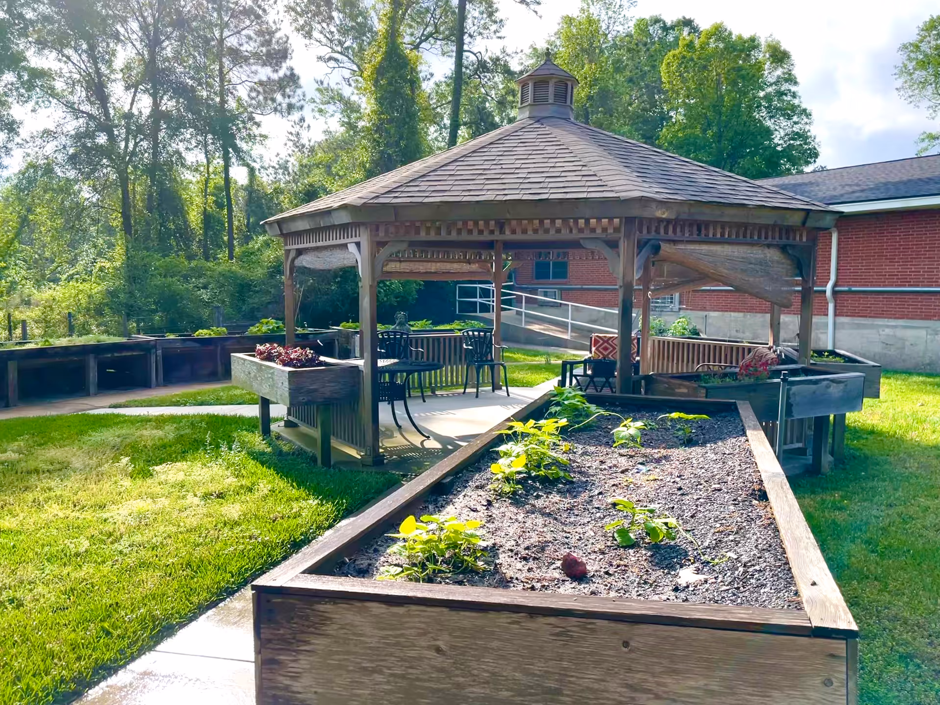 Outdoor garden area at Shady Acres Health & Rehabilitation featuring a wooden gazebo with seating and tables, surrounded by raised garden beds with plants and greenery, and a red brick building in the background.