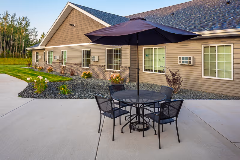 Outdoor patio area at Golden Horizons Of Sandstone featuring a round metal table with four matching chairs and a large dark umbrella. The patio is adjacent to a single-story building with beige siding, brick accents, multiple windows, and air conditioning units. There are landscaped areas with rocks, flowers, and shrubs along the building, and a grassy area with trees in the background.