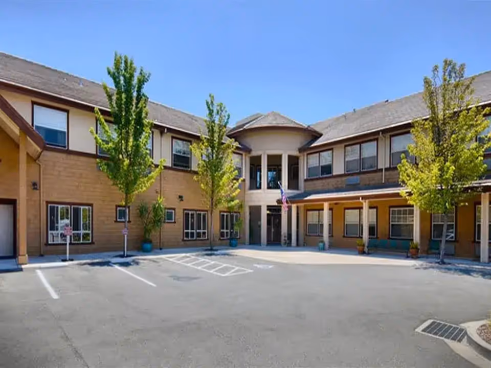 Front exterior of a two-story assisted living building with a covered entrance, small trees, and an empty parking area.