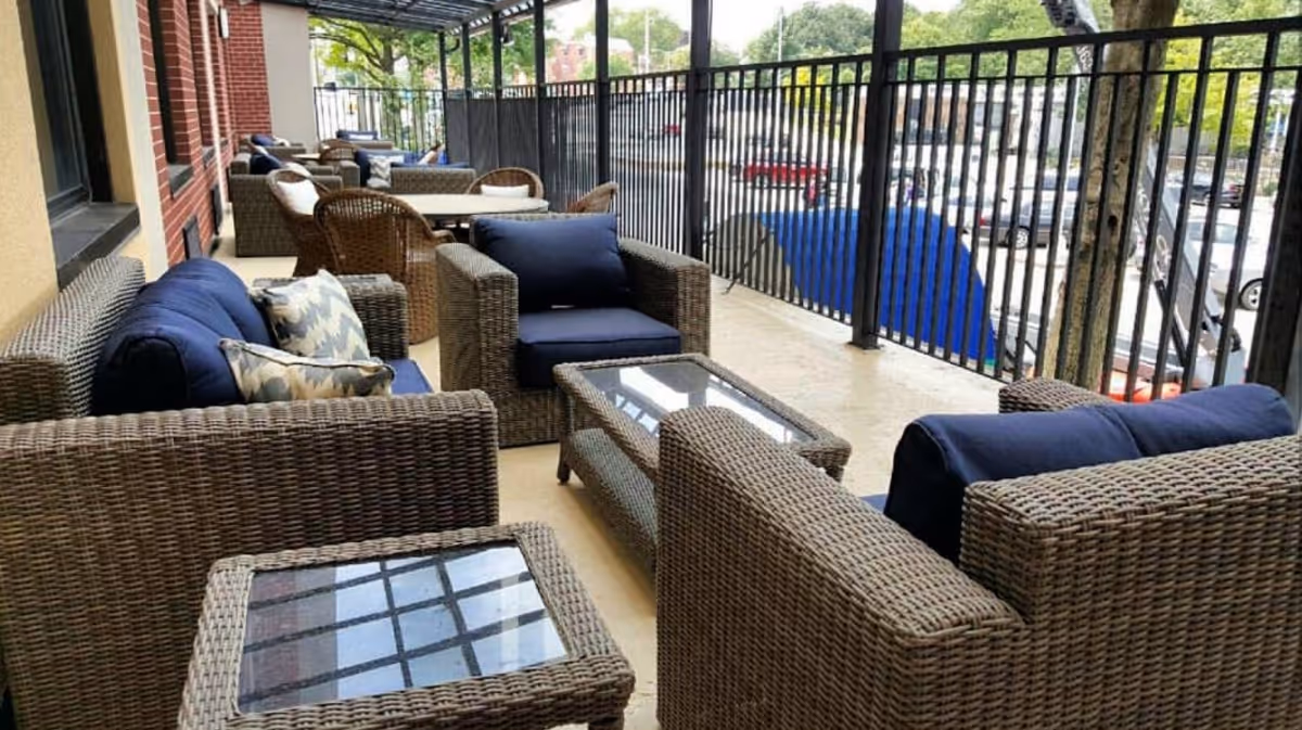 Outdoor patio area with wicker furniture including sofas, armchairs, and glass-top coffee tables. The seating has blue cushions and patterned pillows. The patio is enclosed with a black metal railing and overlooks a parking lot with cars and trees in the background.