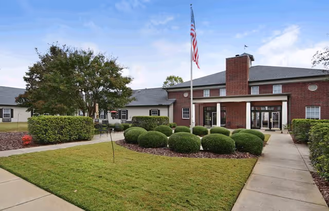 Exterior view of a senior living facility with a brick building, manicured bushes, a flagpole with an American flag, and a paved walkway leading to the entrance under a partly cloudy sky.