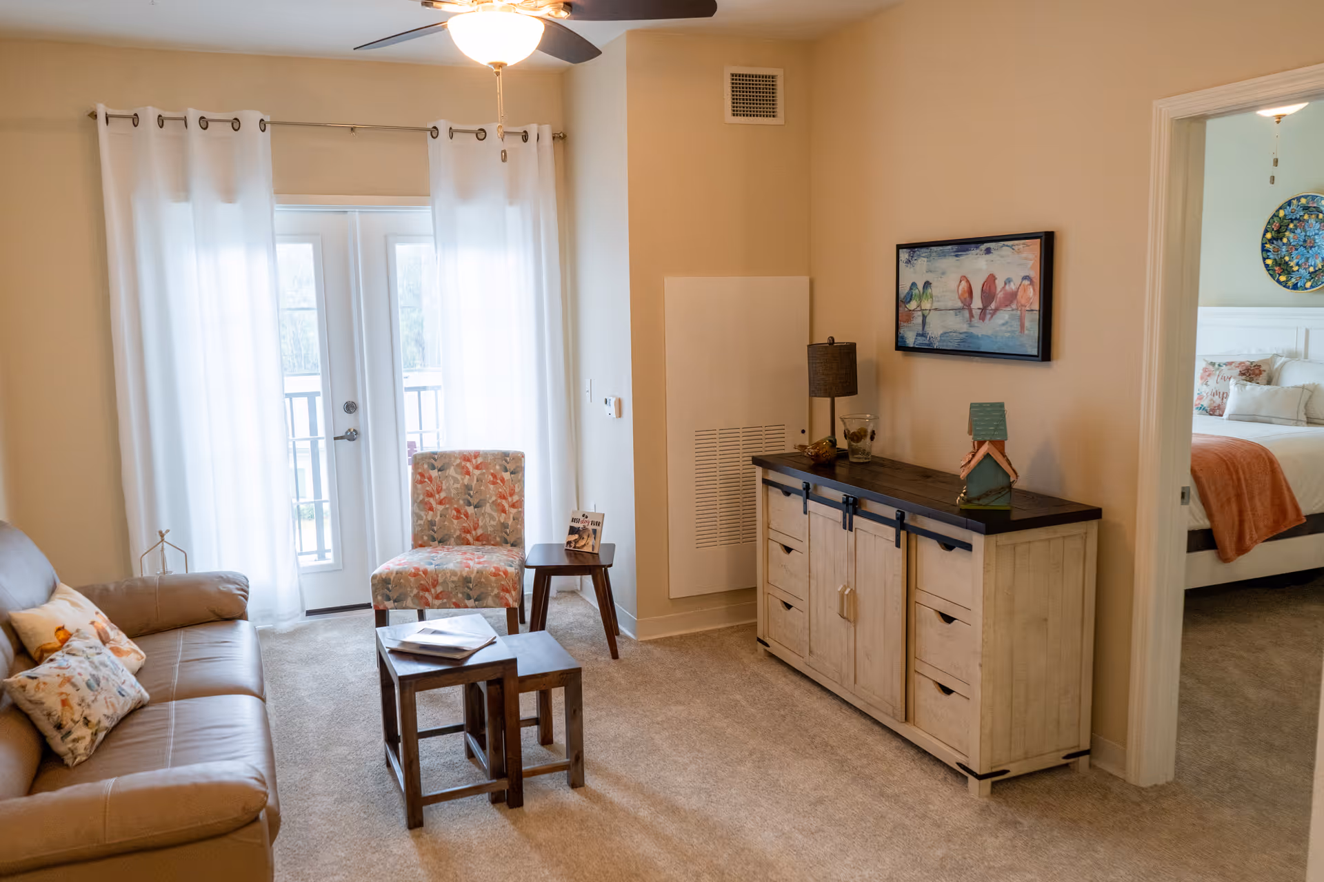 A cozy living room with a beige leather sofa adorned with decorative pillows, a floral patterned chair, and two nested wooden tables. White curtains cover glass doors leading outside. A wooden sideboard with a dark top holds a lamp, decorative items, and a framed painting of colorful birds on the wall above. An adjacent doorway reveals a bedroom with a white bed, floral pillows, and a colorful circular wall decoration.
