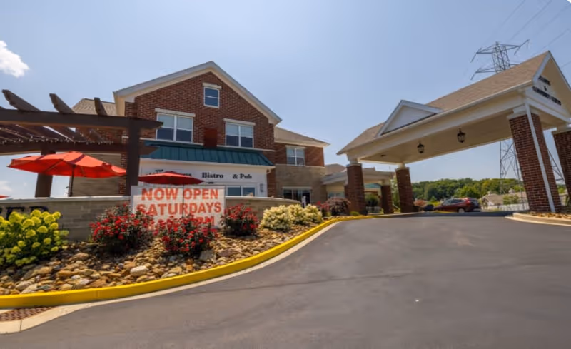 Exterior view of a brick building with a covered entrance driveway and a patio area with red umbrellas and landscaping including bushes and flowers under a clear blue sky.